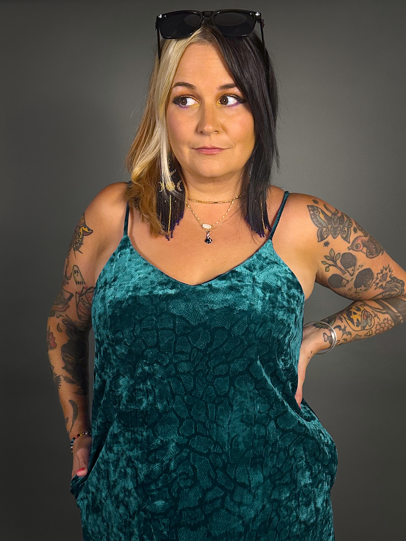 Woman wearing a teal velvet dress with tattoos on arms against a gray background