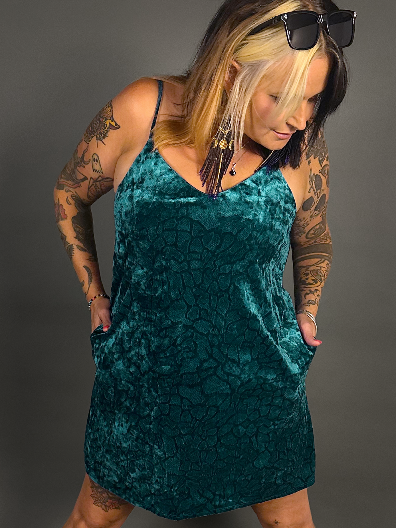 Woman wearing a teal velvet dress with tattoos on arms against a gray background