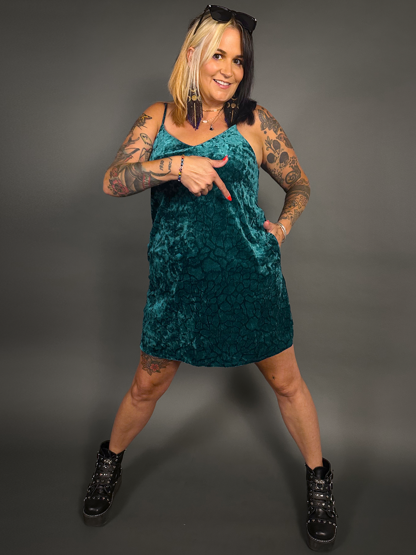 Woman wearing a teal velvet dress pointing at the dress pocket against a gray background