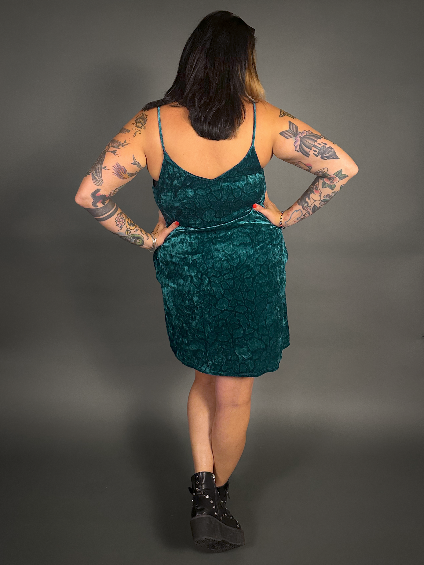 Woman wearing a teal velvet dress with tattoos on arms and legs against a gray background