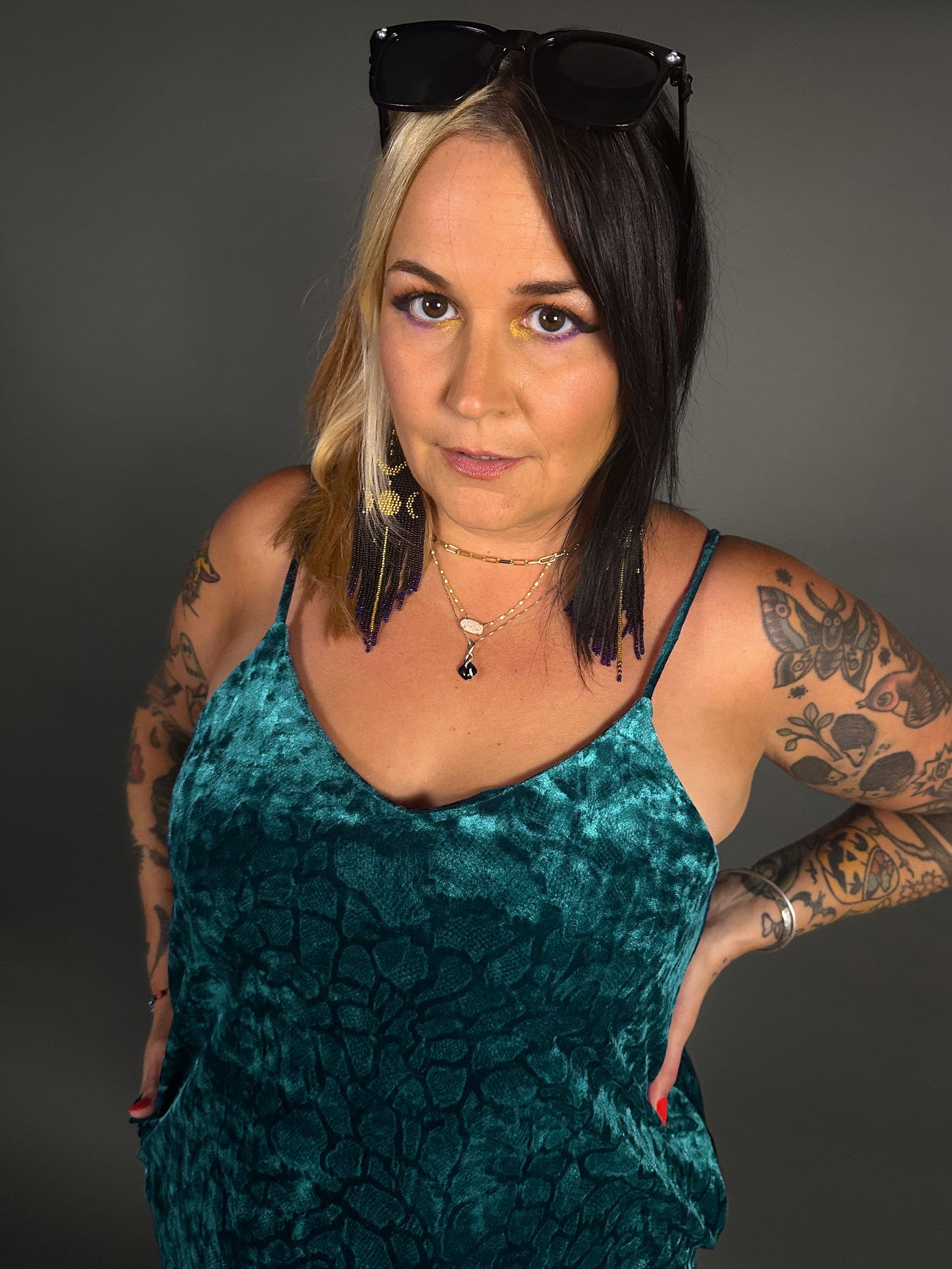 Woman wearing a teal velvet dress with tattoos on arms against a gray background