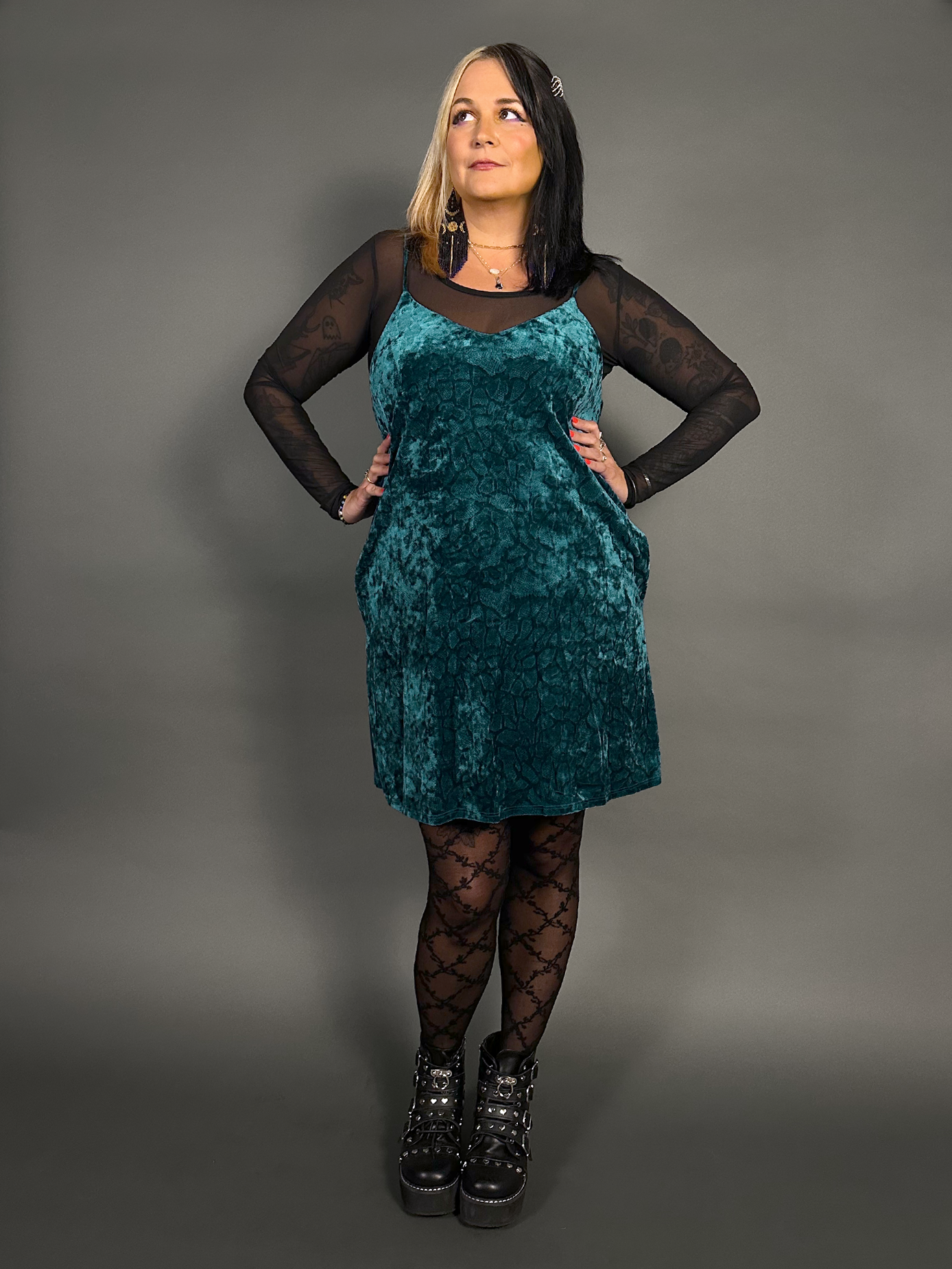 Woman wearing a teal velvet dress with black sheer sleeves and stockings against a gray background
