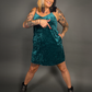 Woman wearing a teal velvet dress pointing at the dress pocket against a gray background
