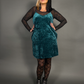 Woman wearing a teal velvet dress with black sheer sleeves and stockings against a gray background