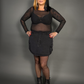 Woman wearing a black sheer top and skirt with black boots against a gray background