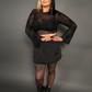 Woman wearing a black sheer top and skirt with black boots on a gray background