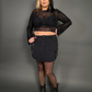 Woman wearing a black sheer top with spiderweb design and skirt against a gray background